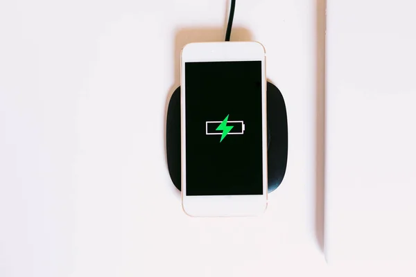 White mobile smart phone with the logo of a battery with green lightning on the screen, charging on a charger base without cable next to the laptop on a white work table