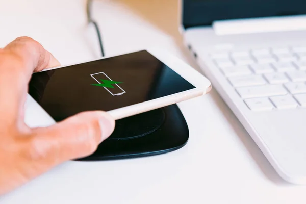 Hand with white mobile smart phone with the logo of a battery with green lightning on the screen, charging on a wireless charger base next to a laptop on a white work table