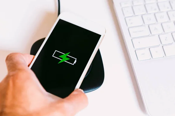 Hand with white mobile smart phone with the logo of a battery with green lightning on the screen, charging on a wireless charger base next to a laptop on a white work table