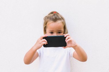 Beyaz t-shirt giyen küçük sarı saçlı kız, cep telefonuna bakıyor, çevirmiş, fotoğraf çekerken.