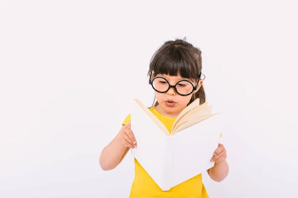 Şaşırmış küçük bir kız, sarı tişört ve yuvarlak siyah gözlüklerle elinde açık bir kitapla beyaz arka planda kitap okuyor.