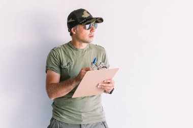 Askeri adam kamuflaj şapkası ve güneş gözlüğü takıyor, bir dosyaya, beyaz bir duvara yazı yazıyor.