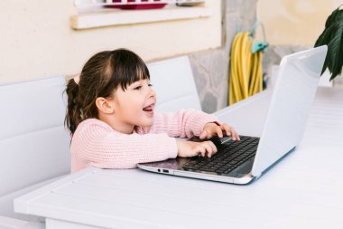 Pembe kazak giyen, dizüstü bilgisayarın önünde oturan, terasta yazan, gülümseyen küçük kızın yan görüntüsü. Çevrimiçi, çocukluk ve teknoloji öğrenimi.