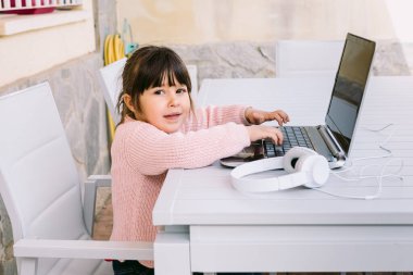 Pembe kazak giyen, dizüstü bilgisayarın önünde oturan, terasta gülümseyen bir kızın yan görüntüsü. Çevrimiçi uzaklık eğitimi, çocukluk ve teknoloji kavramı.