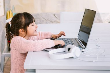 Pembe kazak giyen, dizüstü bilgisayarın önünde oturan, terasta gülümseyen bir kızın yan görüntüsü. Çevrimiçi uzaklık eğitimi, çocukluk ve teknoloji kavramı.