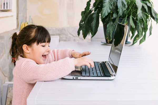 Pembe kazak giyen, dizüstü bilgisayarın önünde oturan, terasta yazan, gülümseyen küçük kızın yan görüntüsü. Çevrimiçi, çocukluk ve teknoloji öğrenimi.