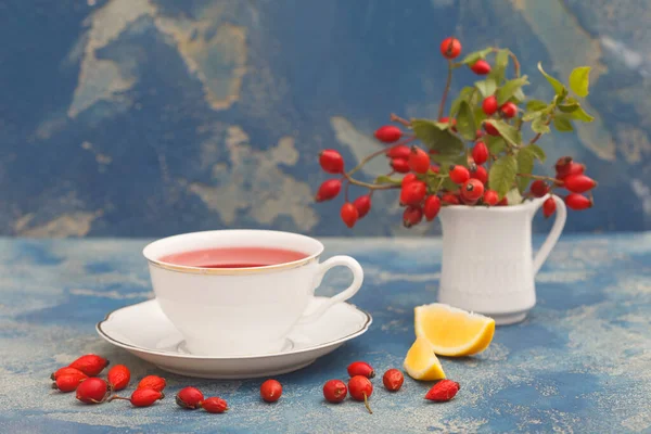 Cup of rosehip herbal tea with rose hip fruits and lemon on blue rustic background. Dogrose autumn or winter healthy tea.