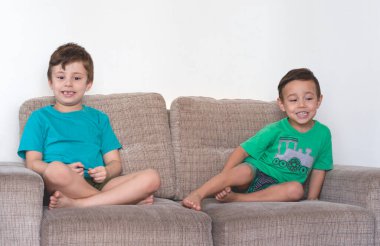 Oturma odasındaki kanepede oturmuş gülümseyen iki küçük kardeşin fotoğrafı.