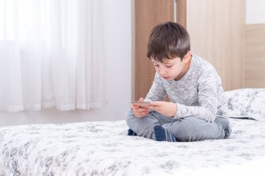 6 yaşındaki küçük bir çocuk akıllı telefon kullanıyor ve oynuyor. Küçük tatlı çocuk yatakta akıllı telefonda oyun oynuyor. Çocuk akıllı telefonuyla video oyununda oynuyor. Sosyal ve teknoloji kavramı.