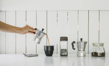 Mokapot kahvesi bardağa dolduruluyor, masada kavrulmuş kahve çekirdekleri, ev içi ya da ofis içi beyaz ahşap arka plan 