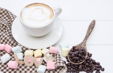 Kahve fincanı beyaz hardal latte sanat ve marşmelovlu tatlı, kavrulmuş kahve çekirdekleri beyaz ahşap.