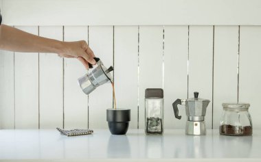 Mokapot kahvesi bardağa dolduruluyor, masada kavrulmuş kahve çekirdekleri, ev içi ya da ofis içi beyaz ahşap arka plan 