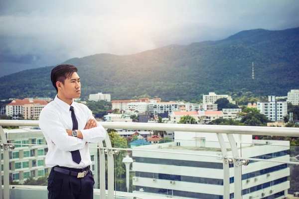 白いシャツを着た若いアジアのビジネスマンがテラスに立ち 腕を組んで街中の風景や山々を眺めます — ストック写真