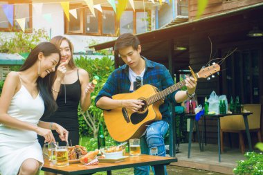 İki Asyalı kadın ve bir erkek gülümsüyor. Eğleniyorlar, gitar çalıyorlar. Grup kutlamaları mutlu bir şekilde kutlanıyor.