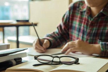 Yakın çekim. Lise öğrencisi elinde kalemle sınavlara giriyor. Günbatımı ışığı arkaplanlı eğitim konsepti için sınıfta yazıyor.