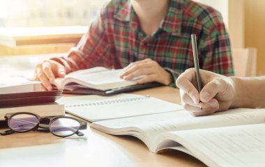 Yakın çekim. Lise öğrencisi elinde kalemle sınavlara giriyor. Günbatımı ışığı arkaplanlı eğitim konsepti için sınıfta yazıyor.