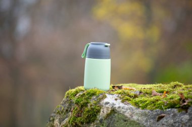 Sıcak kahve ya da çay içeceği olan termos şişesi ormandaki yosun kaplı bir kayanın üzerinde duruyor, seyahat ve seyahat tutkusu konsepti, yiyecek kabı