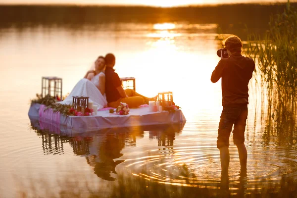 Свадебный фотограф в действии, фотографирует невесту и жениха, сидящих на плоту. Лето, закат — стоковое фото
