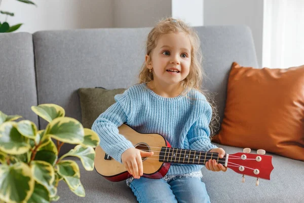 Portrait of cute little girl playing ukulele and singing it at home. 3 years old kid learning guitar. Concept of early childhood education, music hobby, talent, happy preschool leisure and parenting.