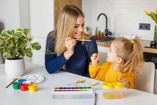 Mother and daughter painting at home. Cute little kid in yellow sweater having fun with parent and paints. Concept of early childhood education, hobby, talent, preschool leisure and parenting.