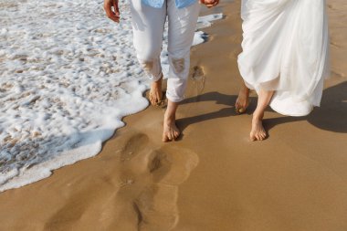 Yeni evlilerin balayı. Beyaz elbiseli çıplak ayaklı gelin ve romantik okyanus sahilinde yürüyen beyaz pantolonlu damat. Genç çiftler altın kumda.