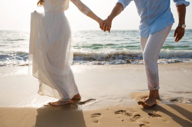Yeni evlilerin balayı. Beyaz elbiseli çıplak ayaklı gelin ve romantik okyanus sahilinde yürüyen beyaz pantolonlu damat. Genç çiftler altın kumda.