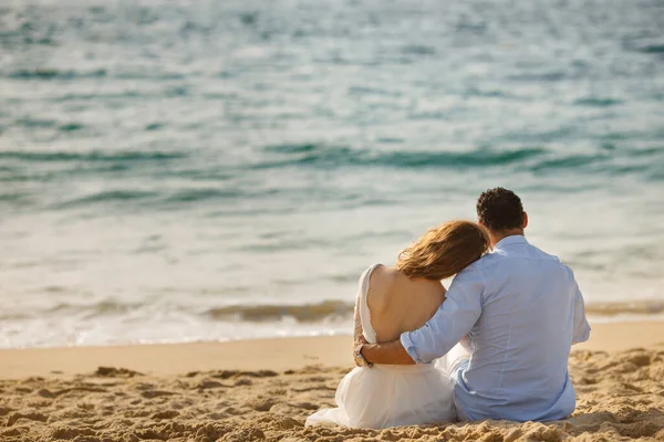 Newlyweds sitting close together at sunset on the beach in the sand, gazing out to sea. Bride and groom embrace each other near ocean and looking on sunset. Concept of happy honeymoon