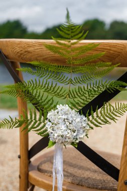 Wooden chair for guests decorated with fern branch, flowers and ribbons in rustic style . Wedding ceremony decor clipart