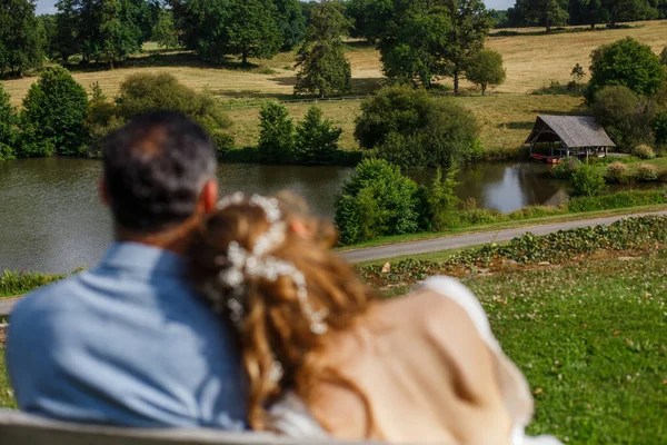 Newlyweds sitting close together on the wooden bench, gazing out to the beautiful landscape view. Bride and groom embrace each other in the park and looking on river. Concept of happy wedding day
