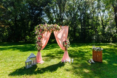 Parktaki düğün merasiminin yapılacağı yer. Tahta dikdörtgen kemer, çiçeklerle süslenmiş beyaz sandalyeler, yeşillik, taç yaprakları, şeftali rengi tül. Sevimli, modaya uygun kırsal dekor