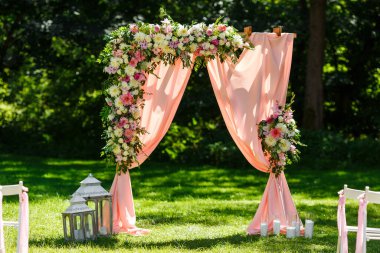 Parktaki düğün merasiminin yapılacağı yer. Tahta dikdörtgen kemer, çiçeklerle süslenmiş beyaz sandalyeler, yeşillik, taç yaprakları, şeftali rengi tül. Sevimli, modaya uygun kırsal dekor