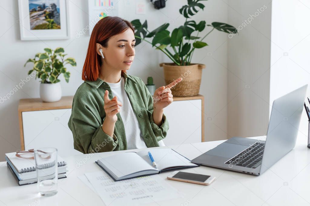 Woman remotely working at home with laptop. Young businesswoman talking via wireless earphones. Concept of online business or communication, freelance, home office and telework for entrepreneur.