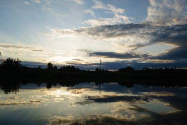Gün batımında ormanın yanında karanlık bir göl, ağaçların yansıması ve güneşli bulutlar.