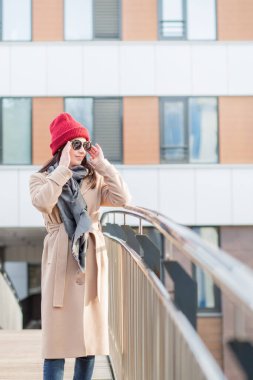 Güneş gözlüklü kadın, kırmızı şapkalı ve paltolu. Sonbahar şehrinde köprüde duruyor.
