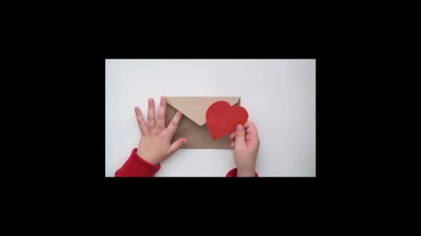 Les mains des enfants mettent du papier rouge dans une enveloppe artisanale. Saint Valentin respectueux de l'environnement — Video