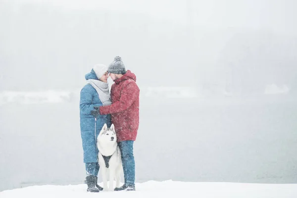 Casal de pé com husky siberiano na margem do rio no inverno. Dia dos namorados. Cão. — Fotografia de Stock