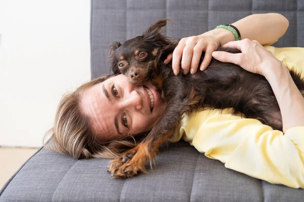 Szczęśliwa blondynka leżąca w zabawny brązowy rosyjski zabawki terrier na kanapie — Zdjęcie stockowe