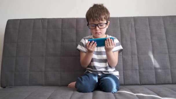 4k 입니다. 안경을 쓴 작고 귀여운 초등 학교 남자 아이가 핸드폰으로 놀고 있습니다. — 비디오