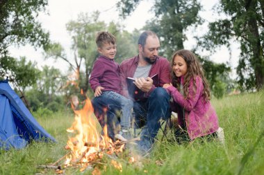 Baba, kız ve oğul ormanda kamp ateşinde oturur ve kitap okurlar.