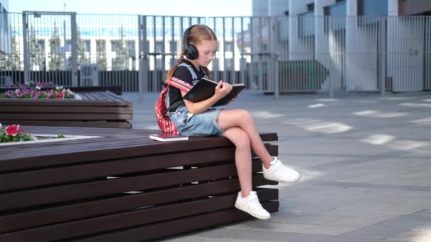 Χαριτωμένο καυκάσιο κορίτσι κοκκινομάλλα σε ακουστικά με σακίδιο κάθονται και να διαβάσετε το βιβλίο — Αρχείο Βίντεο
