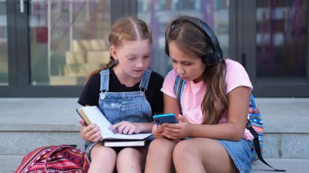 Twee meisjes met rugzak zitten en lezen boek met telefoon — Stockvideo