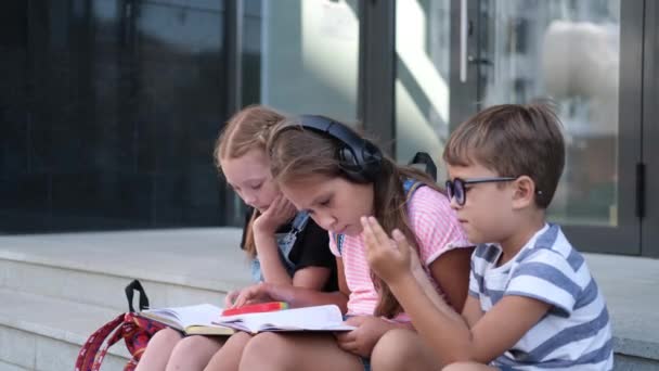 Tres niños con mochila sentarse y leer libro, jugar teléfono — Vídeos de Stock