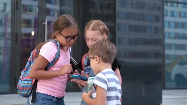 Drie kinderen met rugzak staan samen te praten en te telefoneren. — Stockvideo