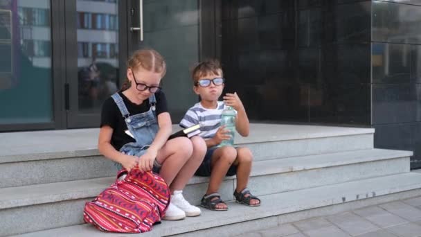 Twee kinderen in brillen met boeken en rugzakken lunchen. — Stockvideo