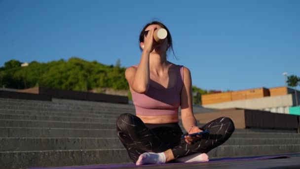 Joven mujer fitness terminar correr, sentarse y beber café y escuchar música — Vídeo de stock