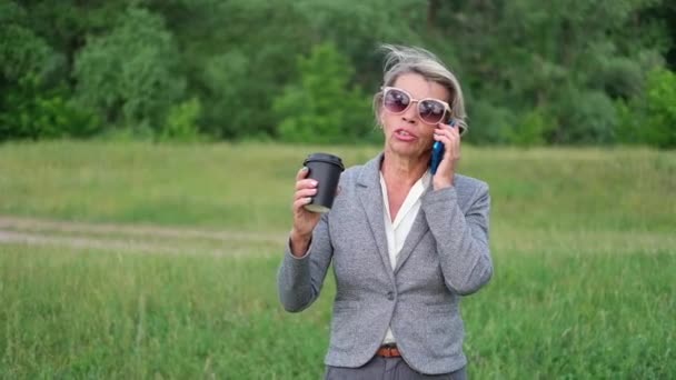 Attractive senior business woman talking on phone, drink coffee, relax in park — Stock Video