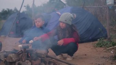 İki çocuk ormanda kamp ateşinde marşmelov kızartıyorlar.