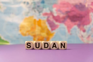 Sudan kelimesi mor bir arkaplan ve coğrafi harita önünde ahşap zarlarla yazılmış.