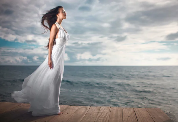 Prachtige vrouw in een witte jurk reputatie op pier dichtbij kliffen. — Stockfoto