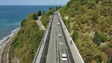 Güzel bir sahil yolu boyunca giden arabaların hava görüntüsü. Deniz kenarındaki kıyı şeridinin manzarası. Köprüden Araba Geçidi Olarak Uçmak. Sochi, Rusya. üst taraf görünümü